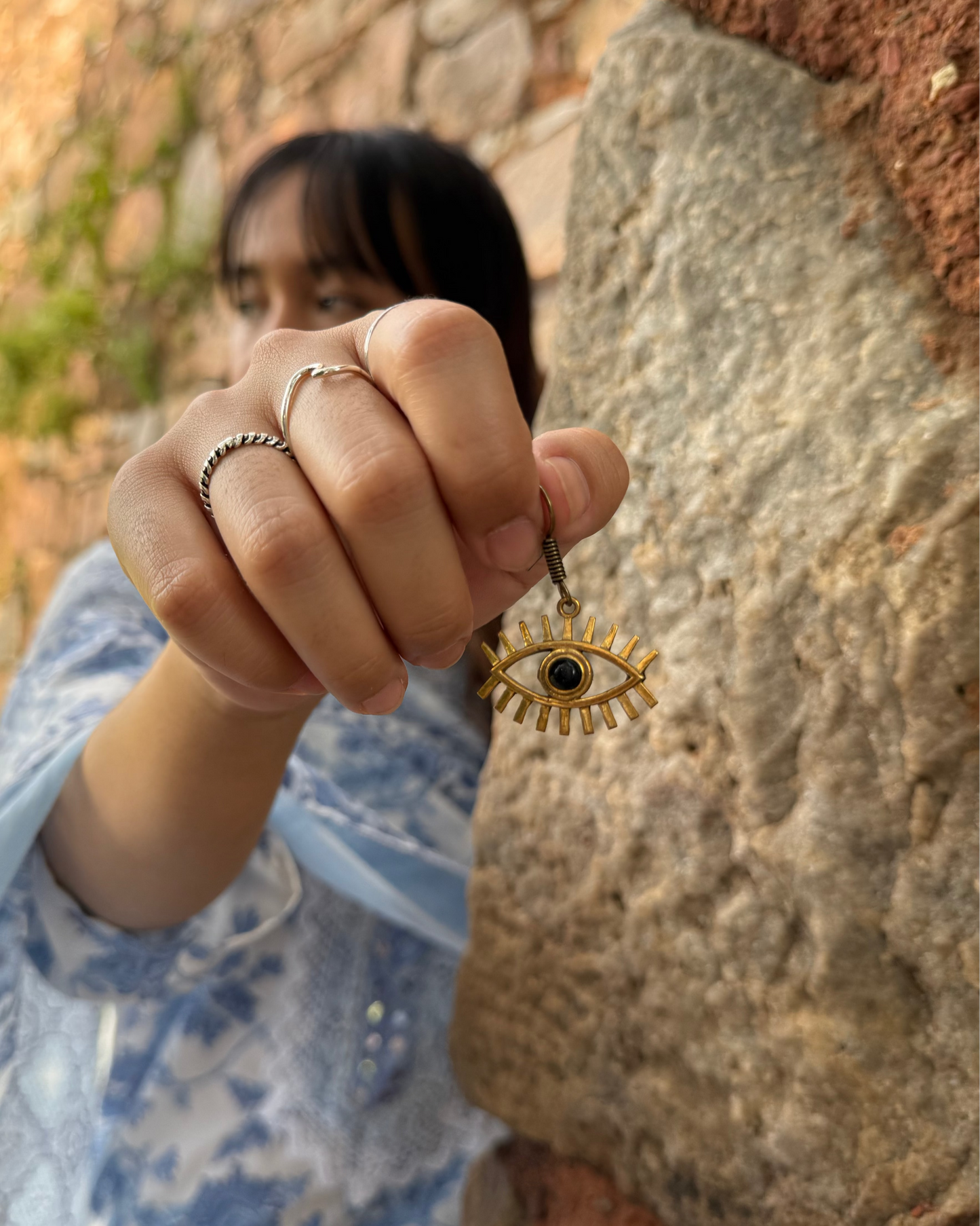 Person holding a gold eye-shaped earring against a stone wall background