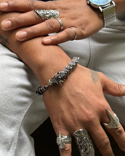Close-up of hands with multiple silver rings and a watch, wearing a plaid shirt and light-colored pants.